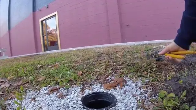 Cleanout pipe with a dark opening set in gravel and grass beside a wall.