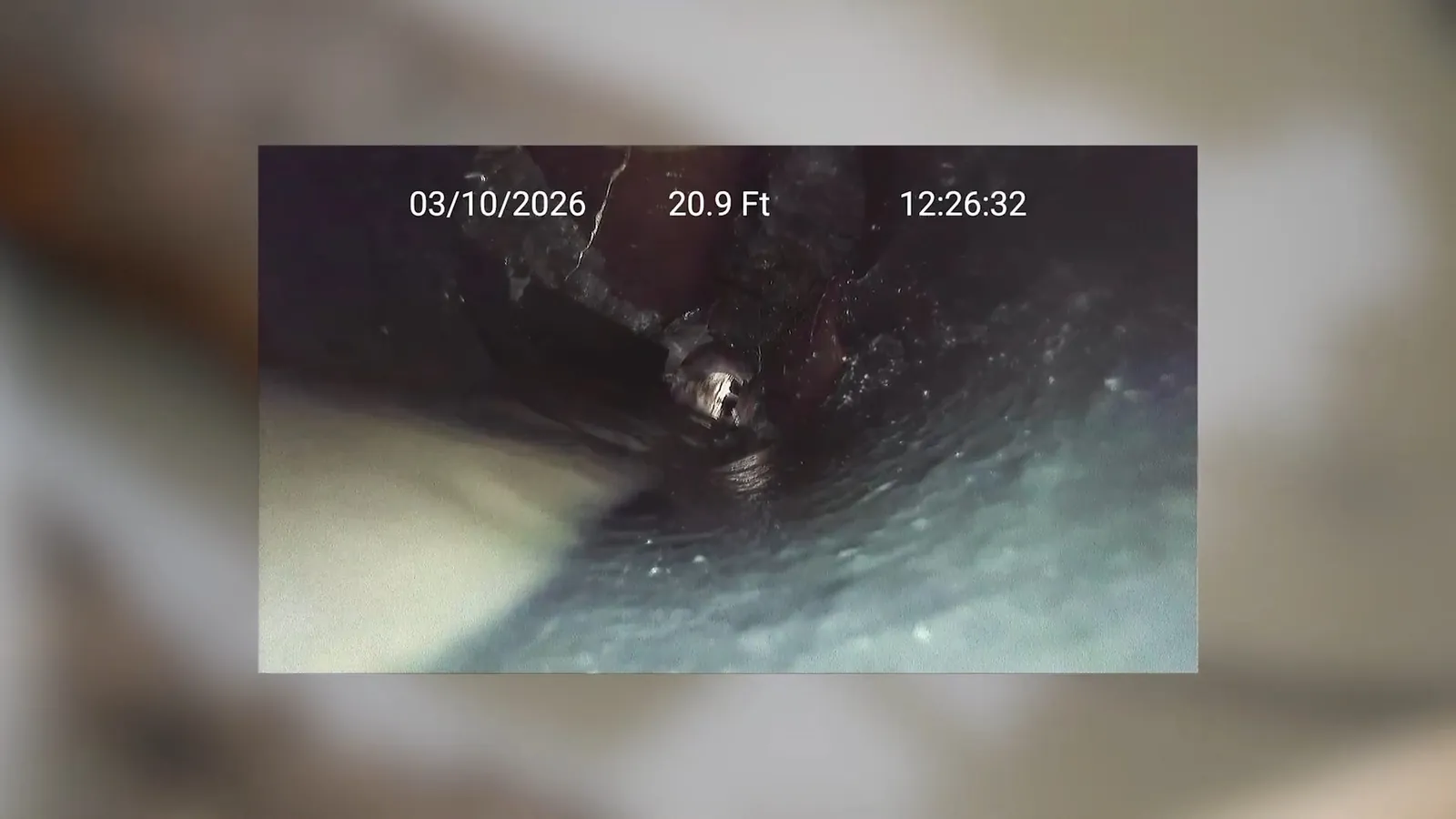 Video frame from a sewer inspection camera showing a pipe interior with a date and depth overlay.