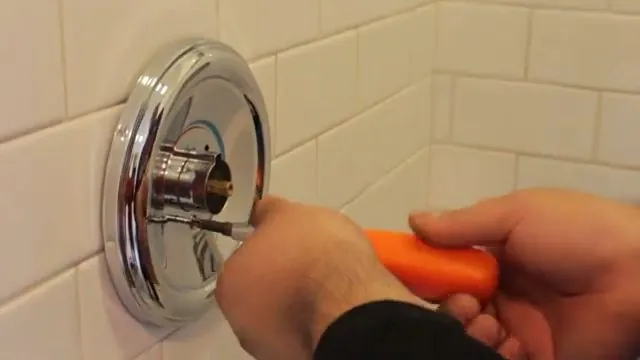 Hands with an orange screwdriver at a chrome shower valve trim plate on a tiled wall.