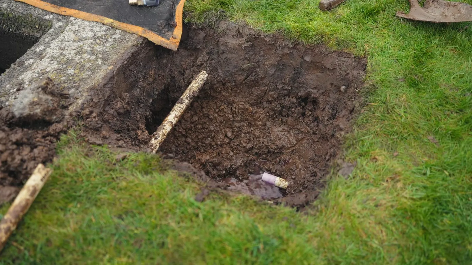 Square access hole dug in a lawn with a length of pipe lying in it.
