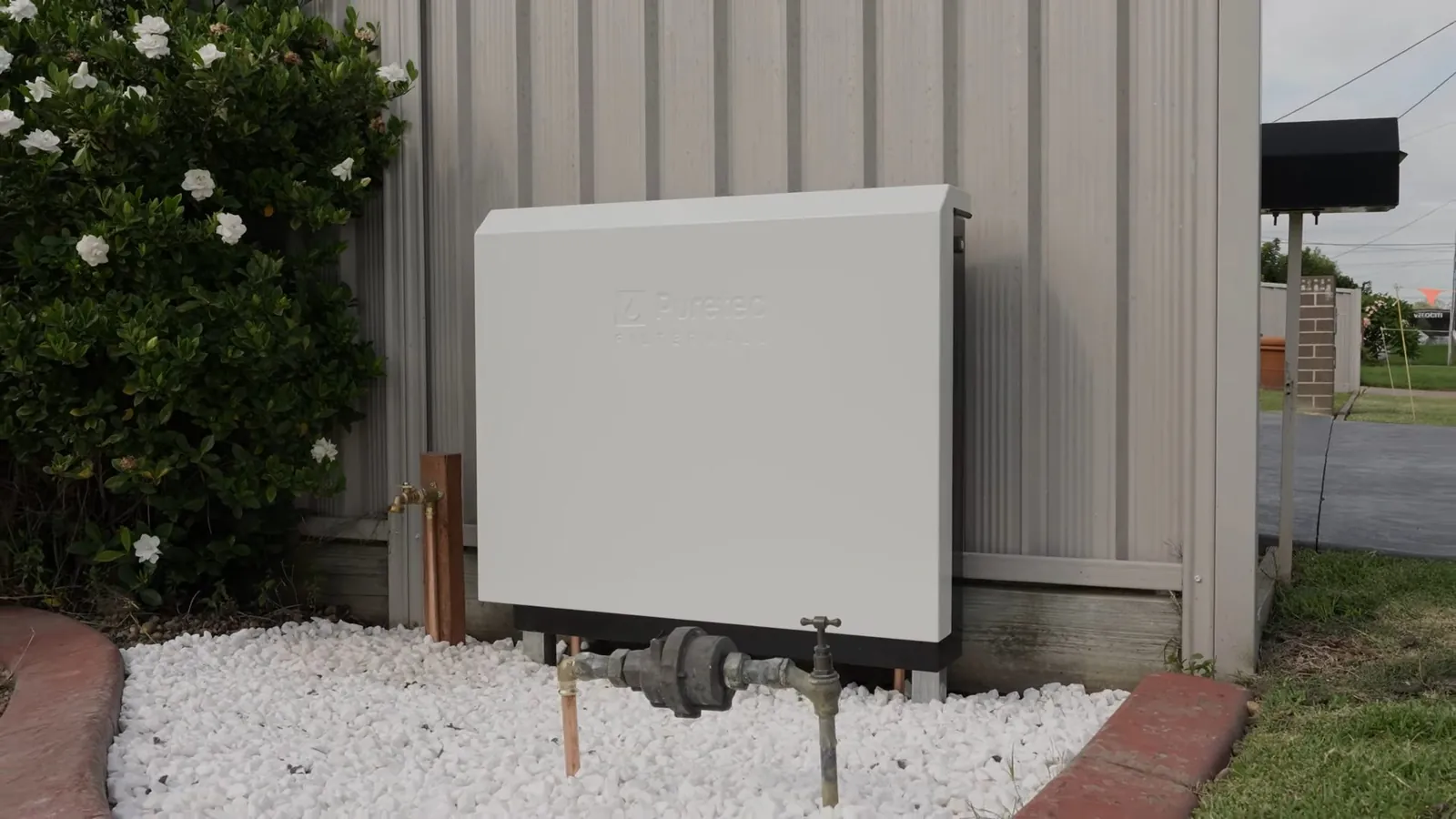 White tankless water heater cabinet on the outside wall of a house above a gravel border.