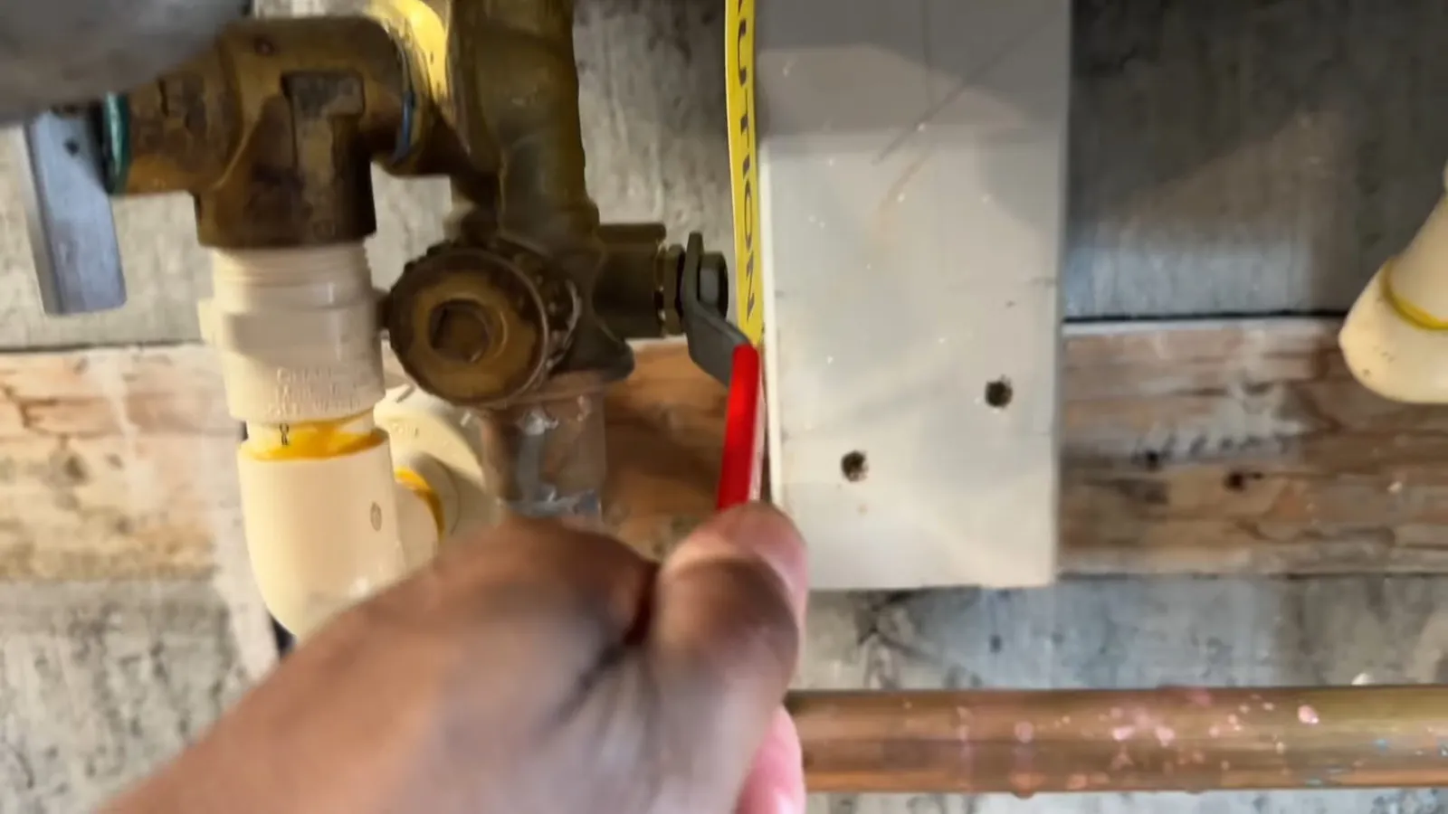 Hand holding a tape measure against brass valves and copper supply pipes on a basement wall.