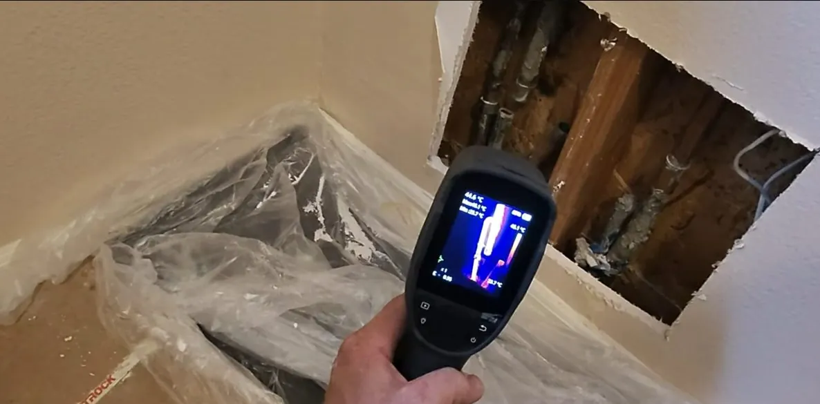 Hand-held thermal camera showing a heat image beside an opened wall cavity with pipes inside.