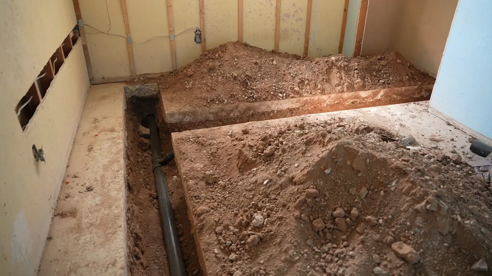 Trench cut through a basement floor with mounded soil on both sides and a dark pipe running along the bottom.