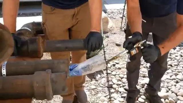 Two workers in work boots handling a liner and rusted pipe sections on gravel.