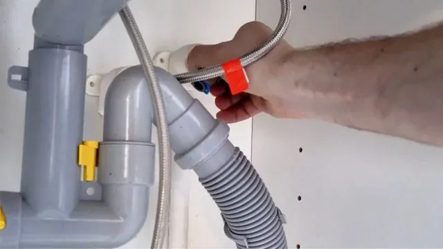 Hand pressing a red clip on a gray plastic P-trap with a ribbed drain hose beside it.