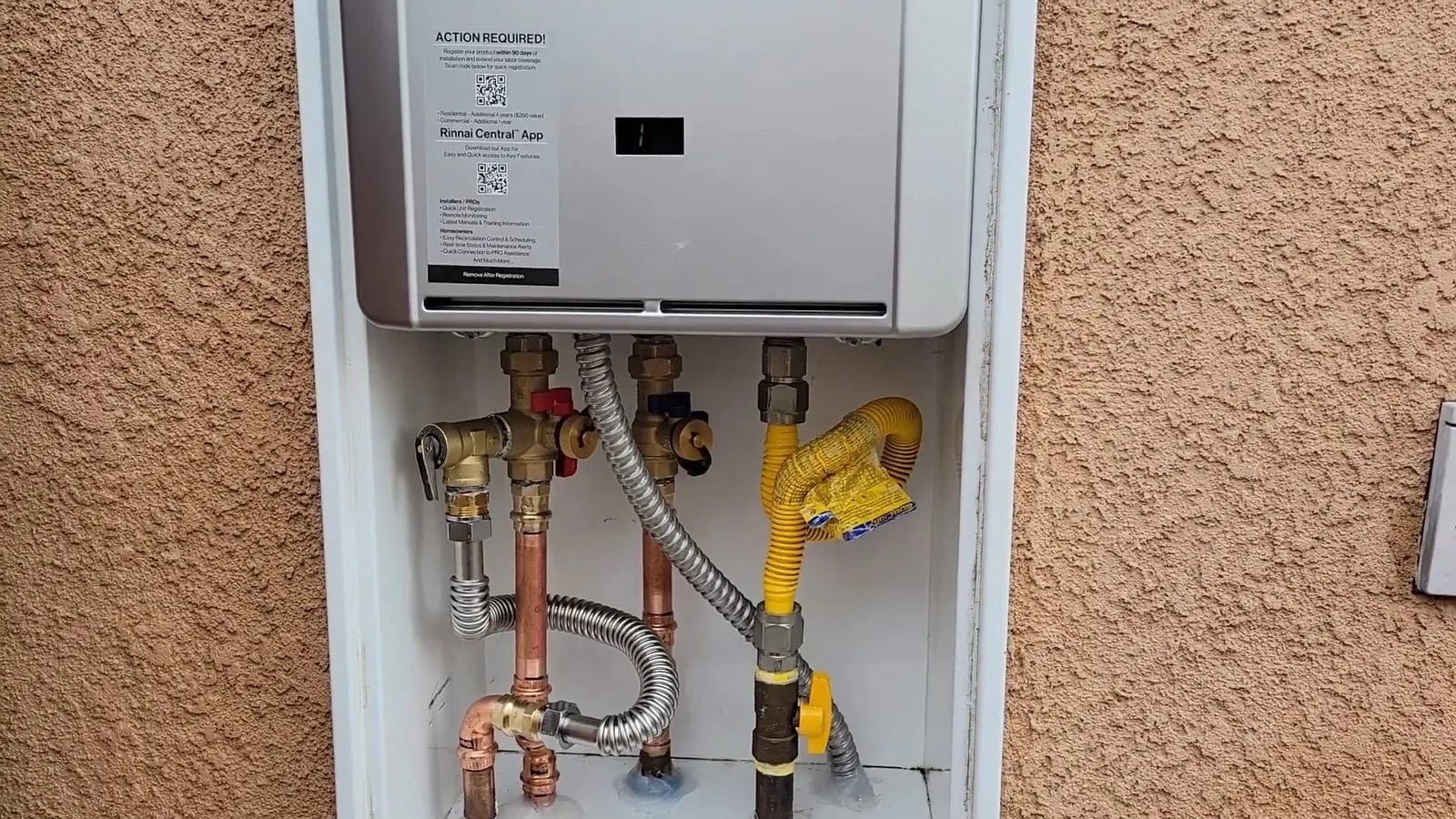 Wall-mounted tankless water heater in a recessed stucco cabinet with isolation valves and a gas line below.