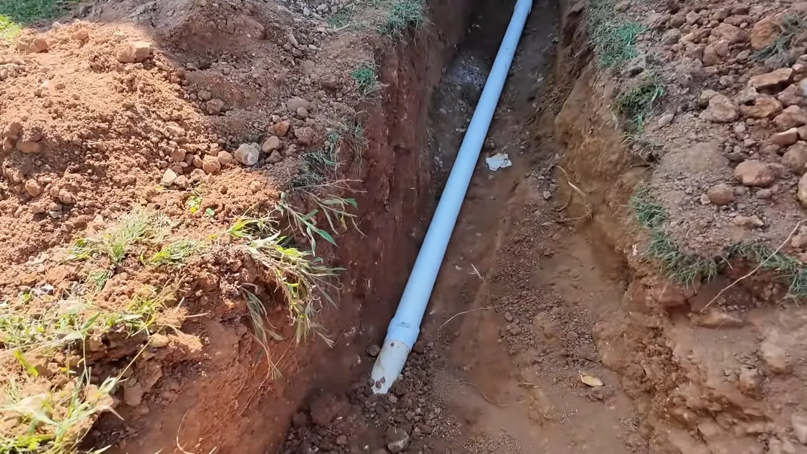 Long white pipe lying in a narrow trench cut through red clay soil.