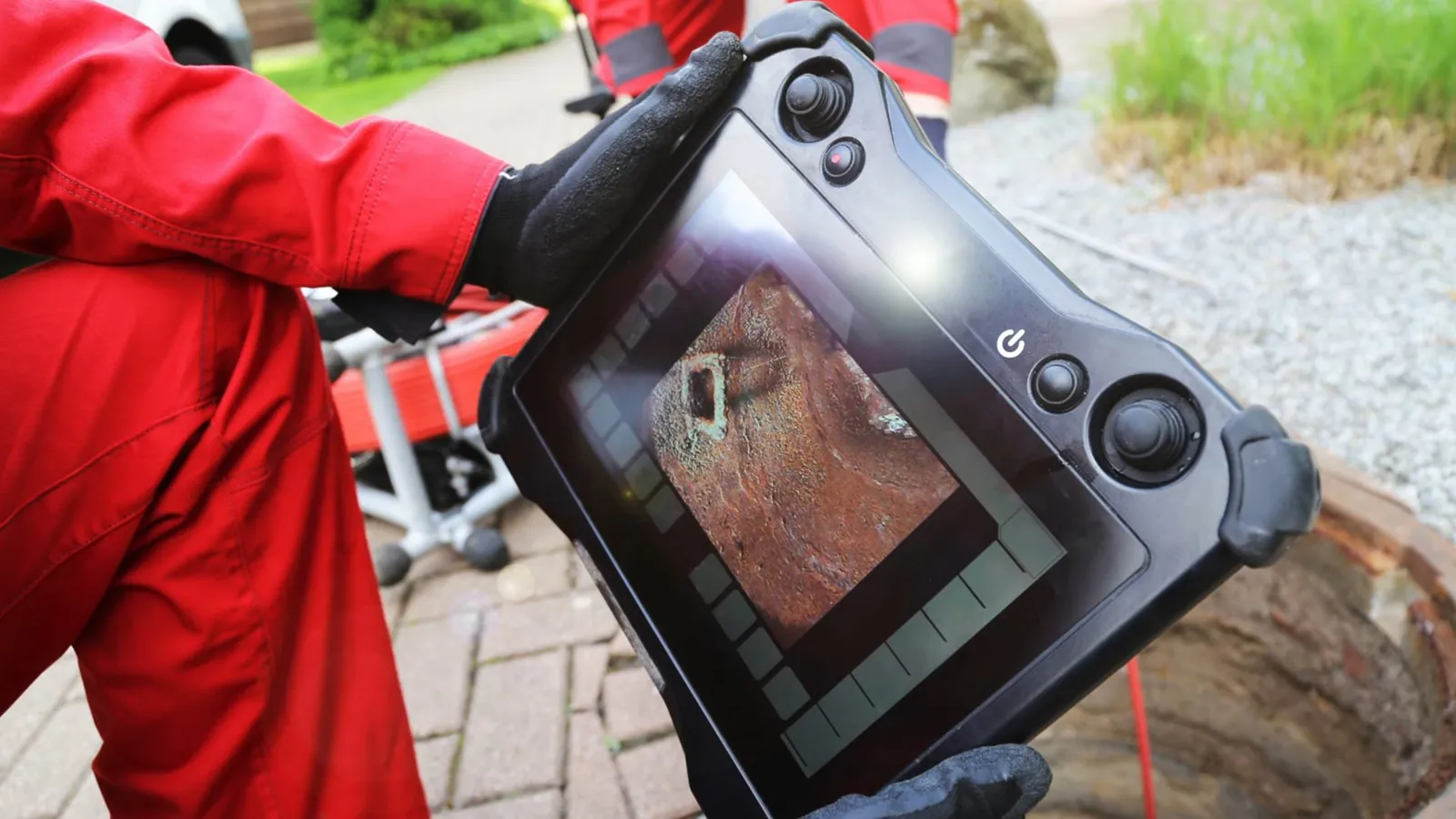 Worker in a red jacket holding a camera monitor beside an open sewer cleanout on a brick path.